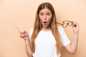 Fototapeta premium Young pretty blonde woman with glasses isolated on beige background surprised and pointing side