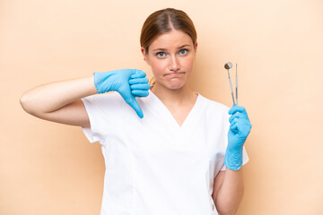 Dentist caucasian woman holding tools isolated on beige background showing thumb down with negative expression