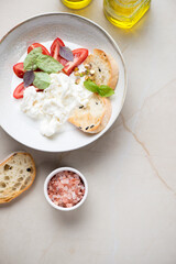 Stracciatella or cheese produced from italian buffalo milk, tomato wedges and ciabatta. Top view on a beige stone background, vertical shot with space