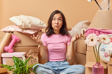 Hesitant dark haired Asian woman sits on floor and shrugs shoulders with doubt doesnt know how to start unpacking cardboard boxes at new home has much work to do during relocation day. Moving in