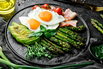 Tasty breakfast. Sunny side up eggs, asparagus, bacon and avocado. On a black stone plate.