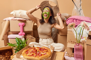 Positive Asian woman applies green facial mask poses crossed legs on floor around cardboard boxes eats delicious pizza relocates to new apartment being in good mood. Ownership and property concept