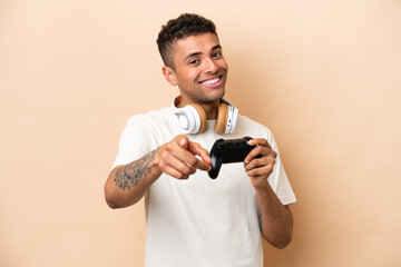 Young Brazilian man playing with a video game controller isolated on beige background pointing front with happy expression © luismolinero