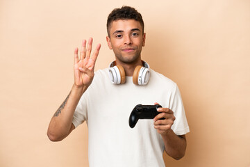 Young Brazilian man playing with a video game controller isolated on beige background happy and counting four with fingers