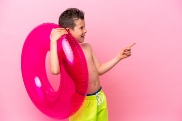 Little caucasian boy holding a inflatable donut isolated on pink background pointing finger to the side and presenting a product