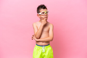 Little caucasian boy wearing a diving goggles isolated on pink background having doubts