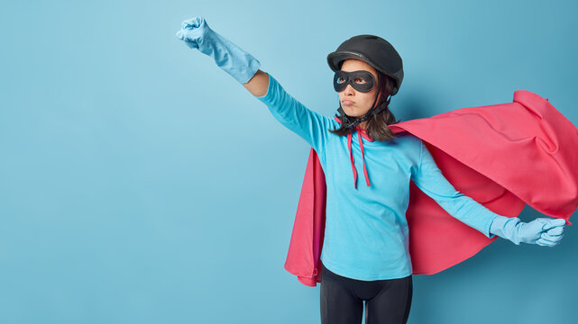 Horizontal shot of brave female superhero ready to save world makes flying gesture clenches fists wears helmet rubber gloves pink cloak poses against blue background blank space for your promo