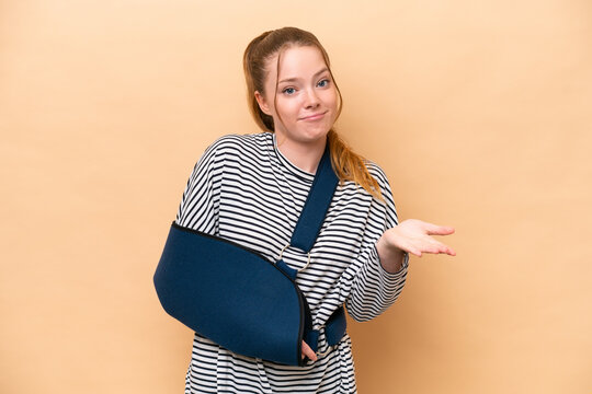 Young caucasian girl with broken arm and wearing a sling isolated on beige background making doubts gesture while lifting the shoulders