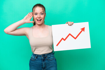 Young caucasian woman isolated on green background holding a sign with a growing statistics arrow symbol with surprised expression
