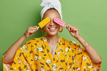 Horizontal shot of cheerful young woman covers eyes with delicious ice cream smiles broadly dressed in yellow robe white soft towel on head feels very happy isolated over bright green background