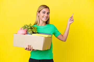 Young Uruguayan girl making a move while picking up a box full of things isolated on yellow background pointing back