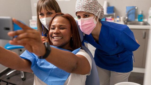 A black woman patient, takes a happy selfie photo with the smartphone side by side the dentist and dental hygienist at the dental clinic