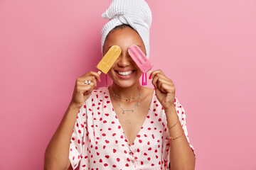 Summer time and junk food concept. Positive young woman covers eyes with two ice creams smiles broadly enjoys favorite dessert dressed in domestic clothes poses against pink studio background.