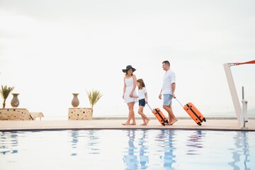 Young couple going to hotel through swimming pool upon arrival, looking for room, holding suitcases