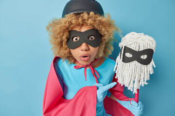 Photo of surprised curly woman pretends being superhero points at mop has shocked expression does household duties suggests to clean your house isolated over blue background. Cleaning service