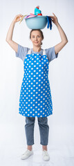 european housewife in a blue kitchen apron, jeans and a t-shirt on a white background, full length portrait. woman holding basin with detergents on her head