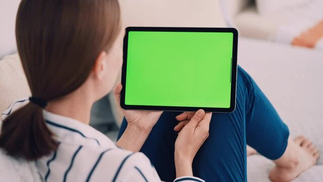 unrecognizable woman sitting on a couch wearing casual shirt and blue trousers using digital tablet with green screen leisure time at home
