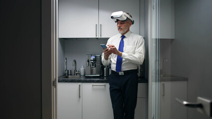 Mature businessman in vr headset using smartphone standing in office kitchen