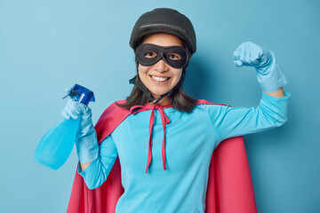 Cheerful active woman pretends being powerful dressed in superhero costume raises arm shows biceps holds bottle of detergent ready for cleaning isolated over blue background. Time for washing