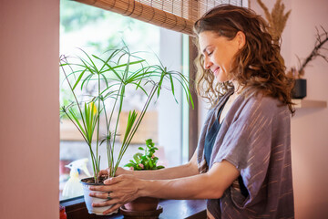 Beautiful happy young woman with indoor plants standing by the window in a country house
