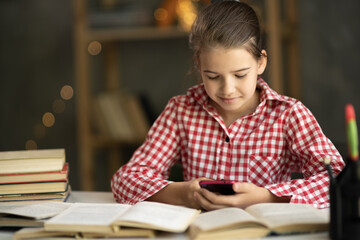Teenage girl school pupil holding smart phone, studying with book. Focused teen student doing homework using mobile app for remote education at home. e-learning