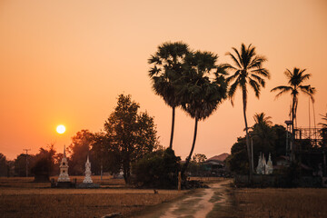 Don Det, Laos - January 19th, 2020 : sunset on the island with silhouettes of stupas and palm trees and a dirt pathway in the foreground