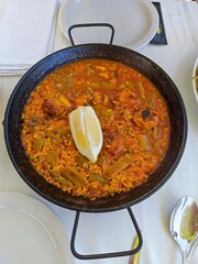 Delicious paella pan with vegetables, meat and a lemon on top served in a restaurant. There are no people or trademarks in the shot.