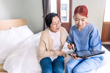 Obraz premium Asian woman nurse wearing scrubs report the health status of Senior Asian woman with digital tablet in the bedroom. Caregiver visit at home. Home health care and nursing home concept.