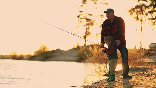 happy granddad and little grandson are catching fish by fishing rod in river in sunset
