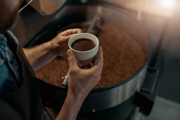 Close up of male barista hands holding cup of coffee on background of roasting coffee machine