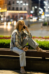 Beautiful young woman talking on phone sitting on the bench at night city street full of neon light.