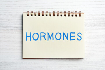 Notebook with word HORMONES on white wooden table, top view