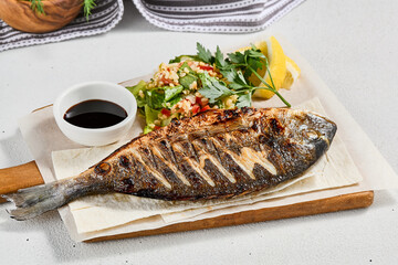 Grilled dorado with garnish on concrete background. Aesthetic composition with bbq fish and vegetables. Healthy food - roasted dorado. Fish dish in minimal style. Whole grilled fish.