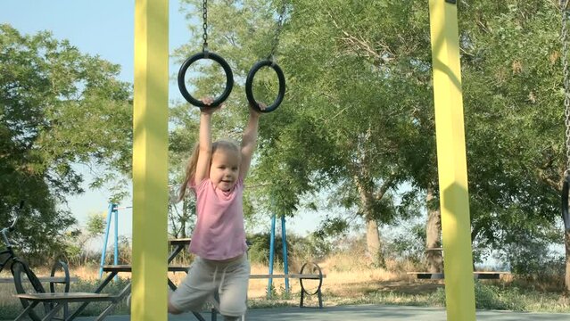 Little girl rides on gymnastic rings on open sports ground on outside. Cute little smiling girl swings on sports gymnastic rings in city park on sun day. Slow motion