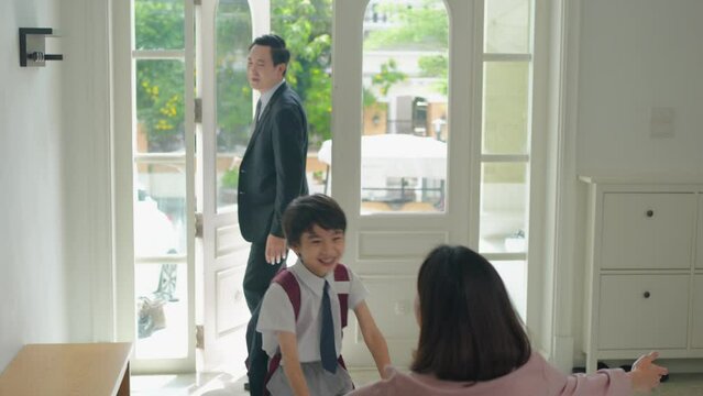 Rack focus shot of an Asian boy in a school uniform with a tie running into the house hugging mother who waiting for him to arrive home safe at the front door, after father picked him up from school.
