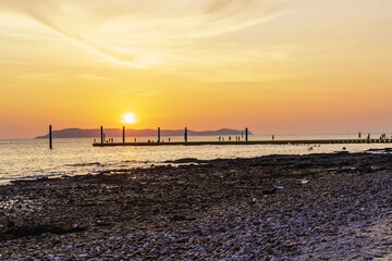 Obraz premium Landscape of colorful sunset shining over bright orange waters of the ocean. Silhouette of tourists walking, relaxing and taking photos sundown on beach of tropical sunset at Koh Larn Beach.