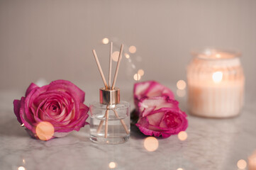 Liquid scented home fragrance in glass bottle and fresh rose flowers over burning candle and glow lights in room. Cozy atmosphere.