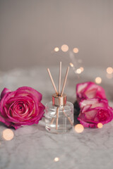 Obraz na płótnie Canvas Liquid scented home fragrance in glass bottle and fresh rose flowers over burning candle and glow lights in room. Cozy atmosphere.