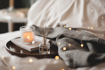 Home liquid fragrance on open paper book and burning scented candle on wooden tray and knit cloth sweater in bed close up. Cozy atmosphere.