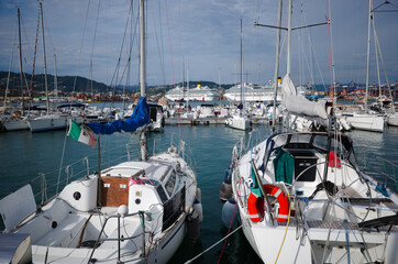 Marina with sailing boats in Porto Mirabello port, La Spezia, Liguria, Italy. Yachts with Italian flag are moored to pier in Mediterranean Sea. Mountains and two large cruise ships in background