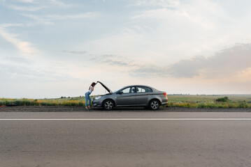 Obraz na płótnie Canvas A young girl stands near a broken-down car in the middle of the highway during sunset and tries to call for help on the phone. Waiting for help. Car service. Car breakdown on road.