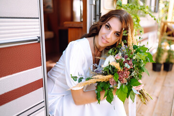 Beautiful stylish bride near trailer. Camping season. Marriage. Happy newlywed woman.