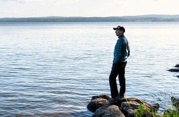 Fototapeta na wymiar young man in a cap looking at the view of the lake or river standing on the stones Enjoying nature landscape