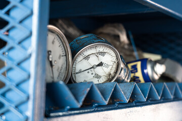 An old pressure gauge or manometer in poor condition (broken glass), which is kept on metal rack in workshop store.