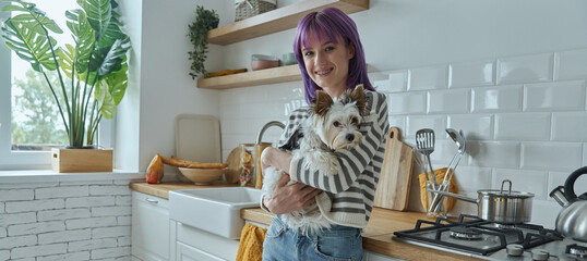 Cheerful young woman with purple hair carrying cute little dog while standing at the kitchen
