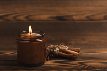 A cozy burning candle in a brown glass jar with cinnamon sticks and pumpkin candles on a brown wood...