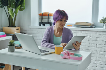 Confident young woman with purple hair working on laptop while sitting at her working place