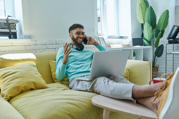 Cheerful African man using laptop and talking on mobile phone while sitting on the couch