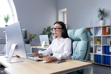 A young beautiful Asian woman is working at a computer in the office. Businesswoman, director, freelancer, manager, sitting in a white shirt and glasses at the desk.