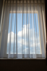 Closed window with a transparent curtain through which you can see the blue sky and clouds. Close-up.